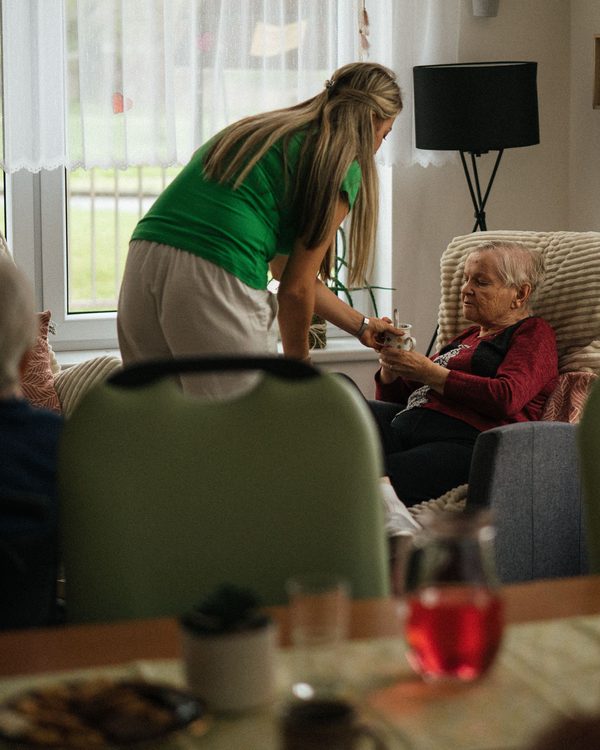 A care worker in a green top leaning down to hold the hands of an older woman sitting in an armchair in a bright care home lounge