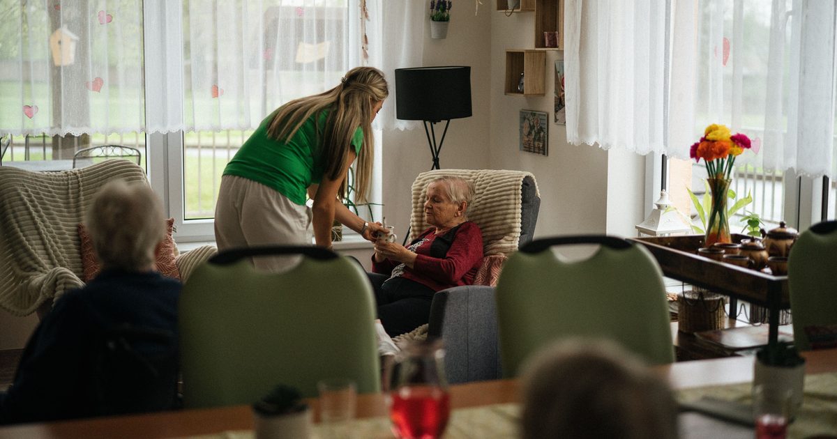 A care worker in a green top leaning down to hold the hands of an older woman sitting in an armchair in a bright care home lounge